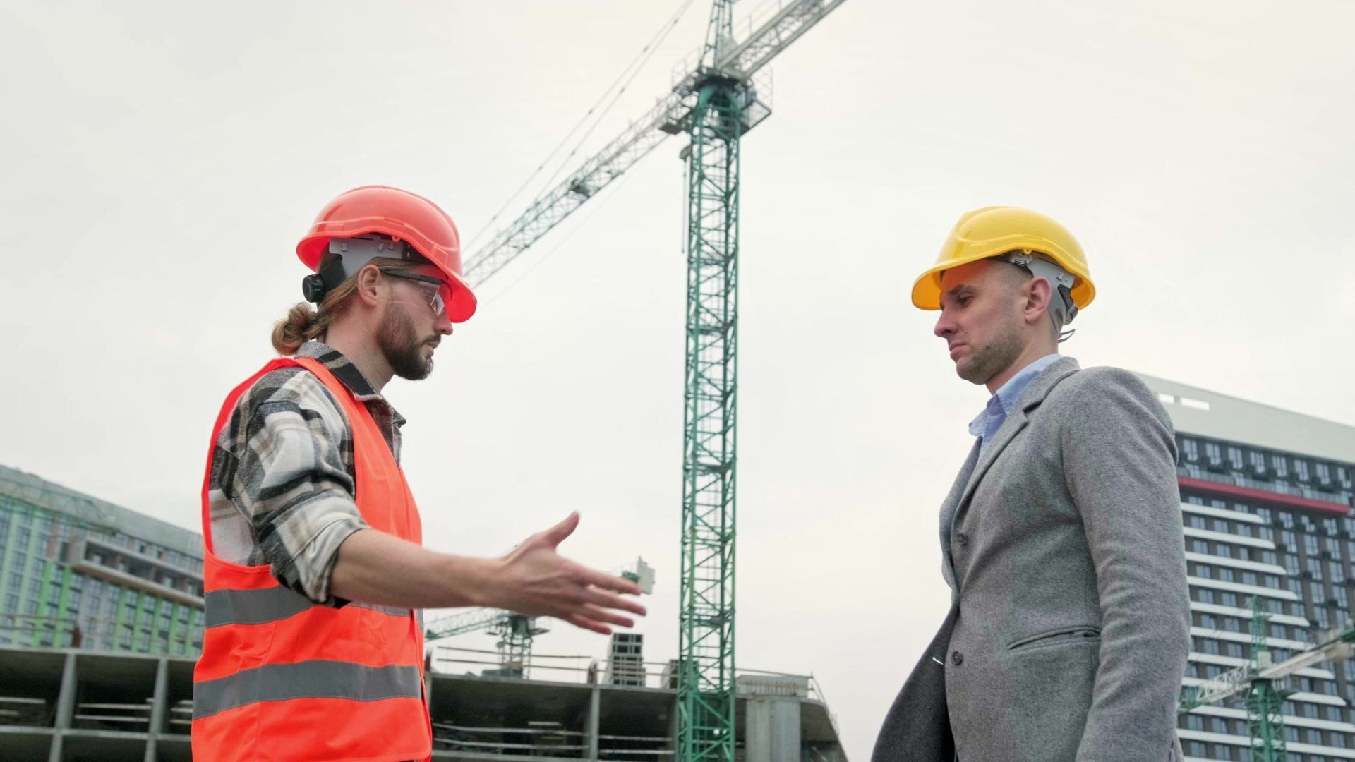 A worker in a hard hat meeting a visitor in a jacket below a tower crane.
