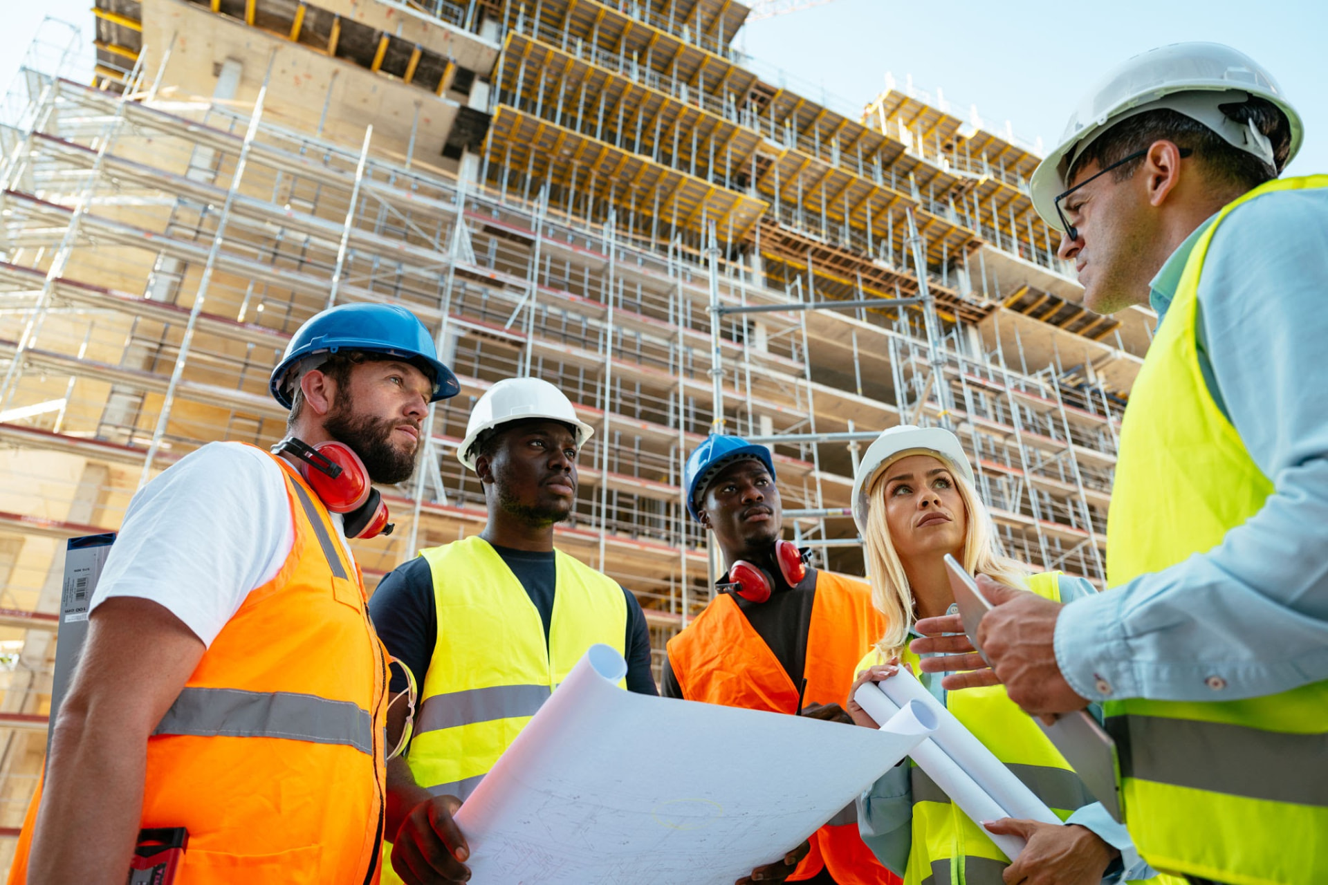 A crew in hard hats and high-visibility vests reviewing rolled plans in front of a scaffolded building.