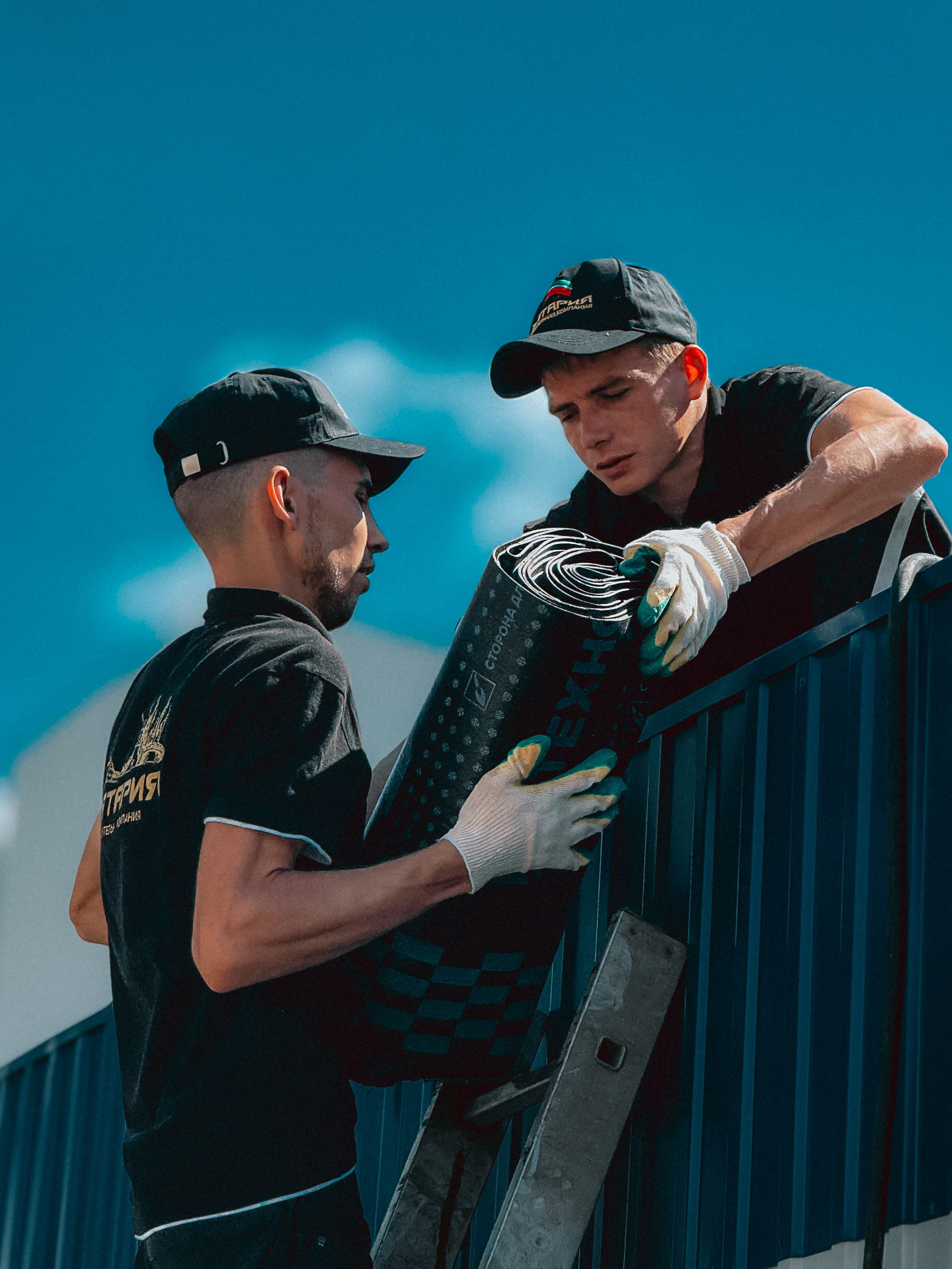 Two roofers passing a roll of waterproofing membrane up a ladder beside a metal-clad building.